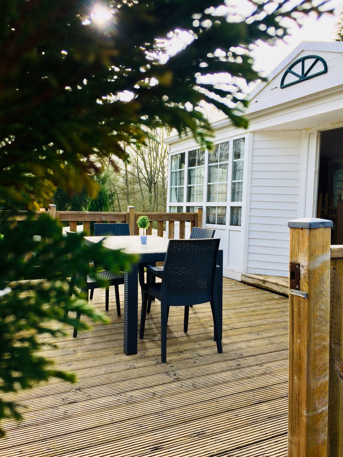 A wooden deck is seen, featuring a round table surrounded by black chairs. Sunlight filters through leaves, creating a natural ambiance. A white structure with large windows is visible in the background, inviting relaxation in a serene setting.