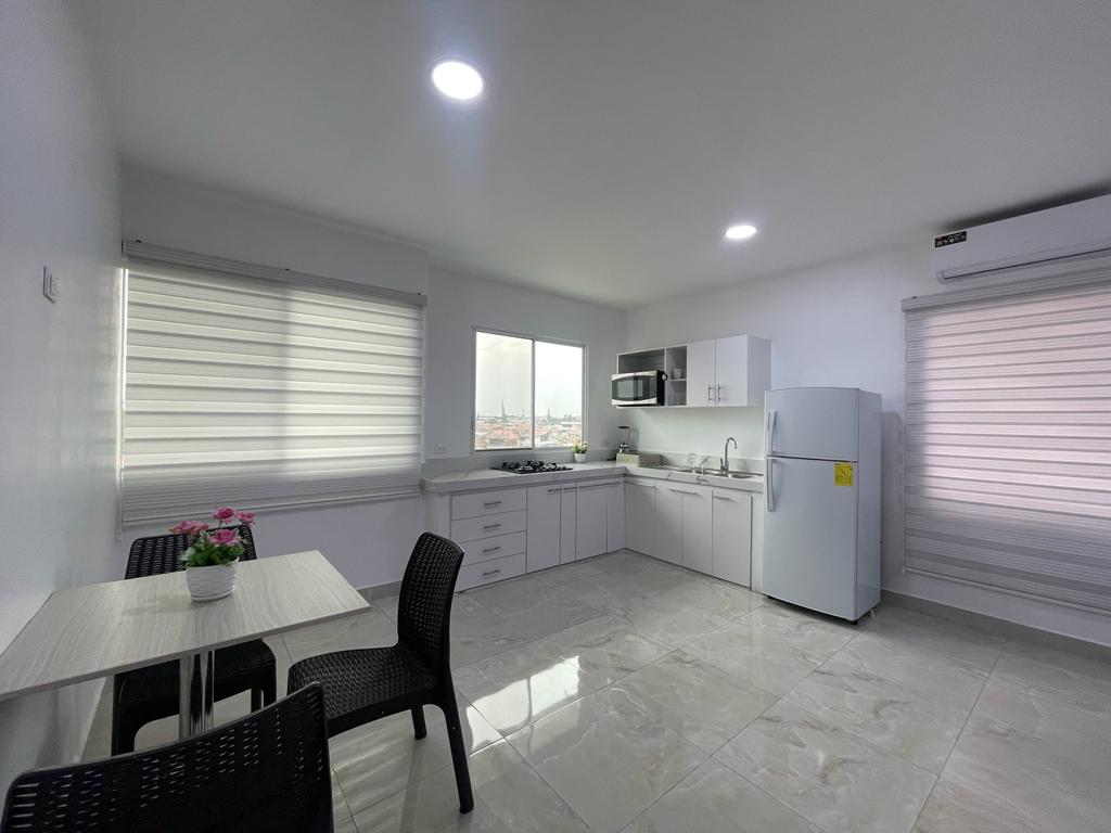 A modern kitchen area is presented with sleek white cabinetry and appliances. Natural light is filtered through two window shades, illuminating the space. A small dining table with two chairs is positioned near a potted plant, complementing the contemporary design.