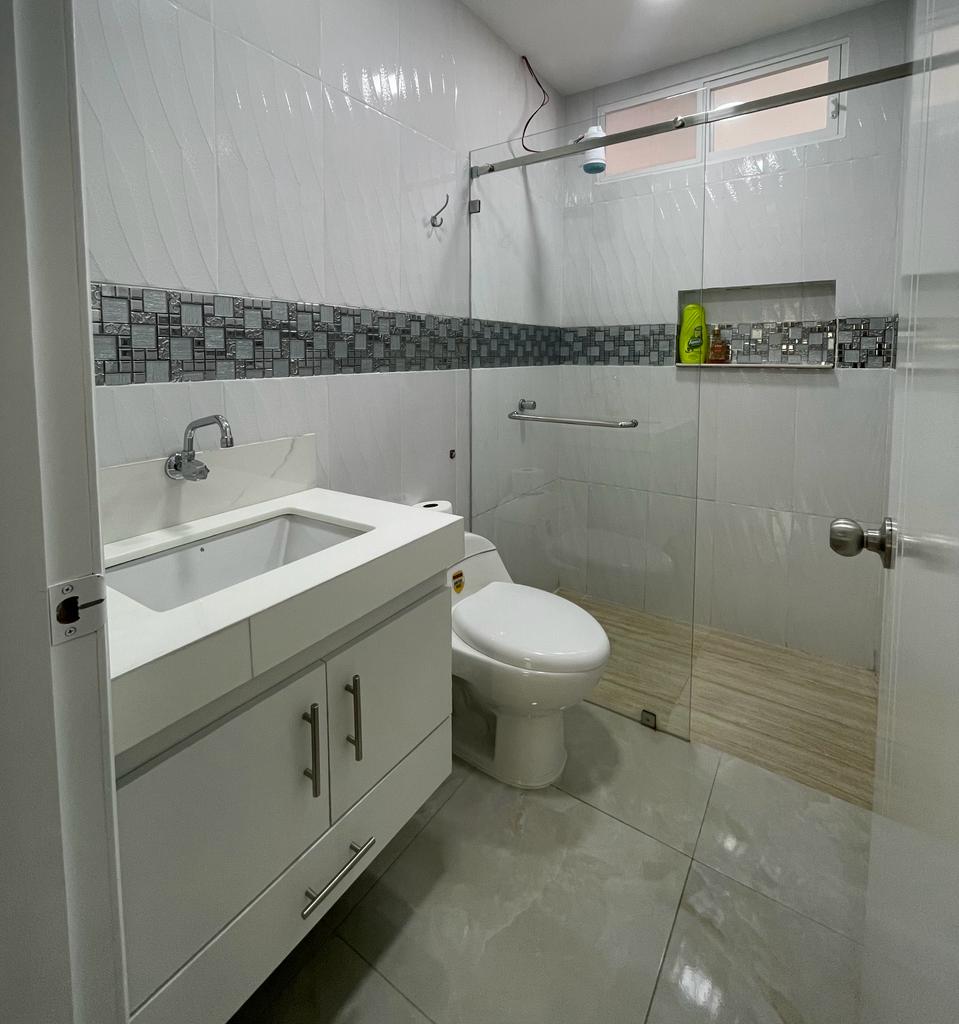 A modern bathroom features a glass-enclosed shower alongside a white toilet and a sleek vanity with storage drawers. The walls are tiled in a clean white and grey design, and a small window allows light to enter the space.