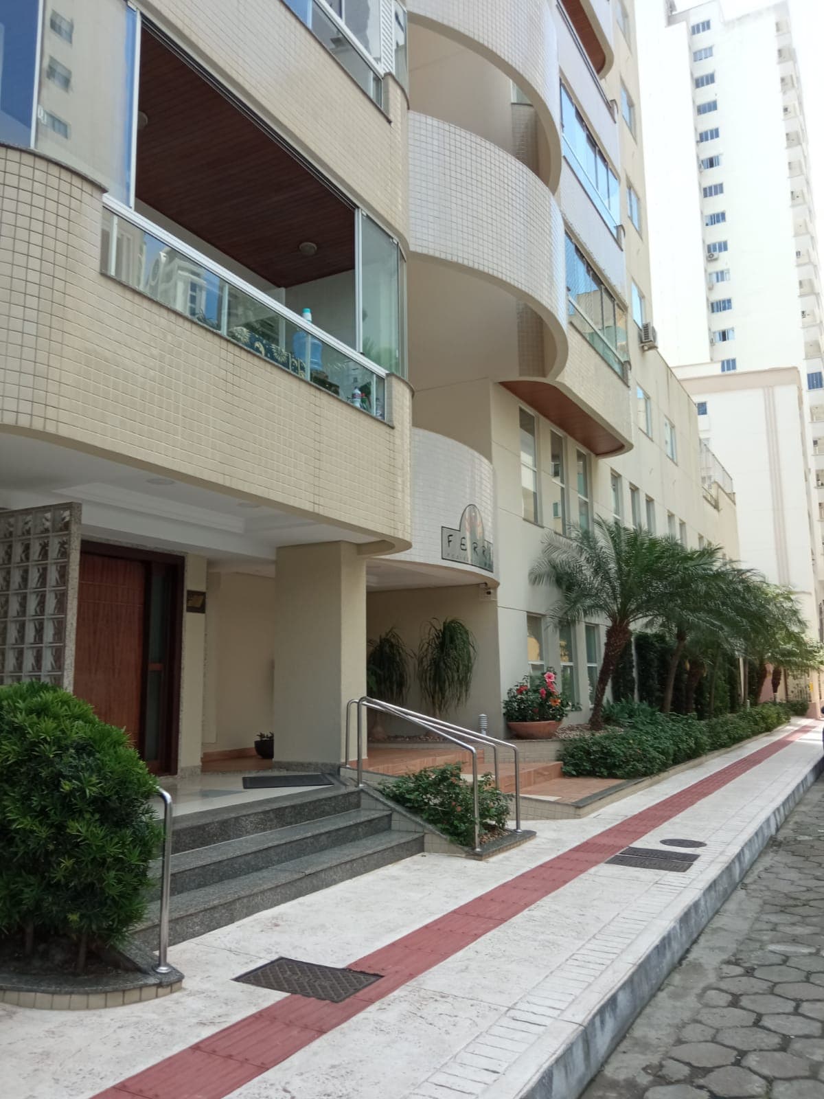 The exterior of a modern building is displayed, featuring a welcoming entrance with polished stone steps. Lush greenery, including palm trees, lines the pathway alongside the building. Large windows offer glimpses of the interior, while a covered area provides shelter at the entrance.