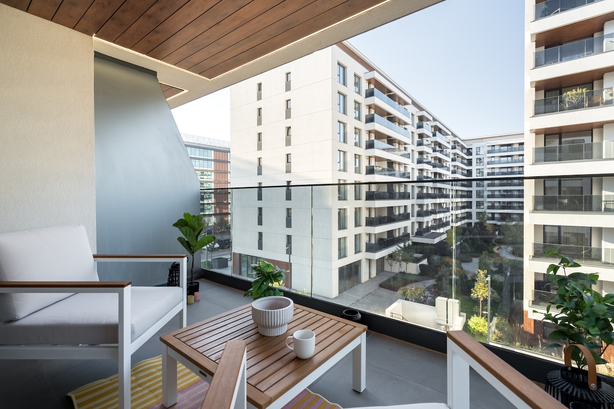 A serene balcony space is presented, featuring comfortable outdoor seating and a wooden table. Lush potted plants adorn the area, with views of the surrounding greenery and modern buildings visible through the glass railing. Natural light fills the space, enhancing the tranquil atmosphere.