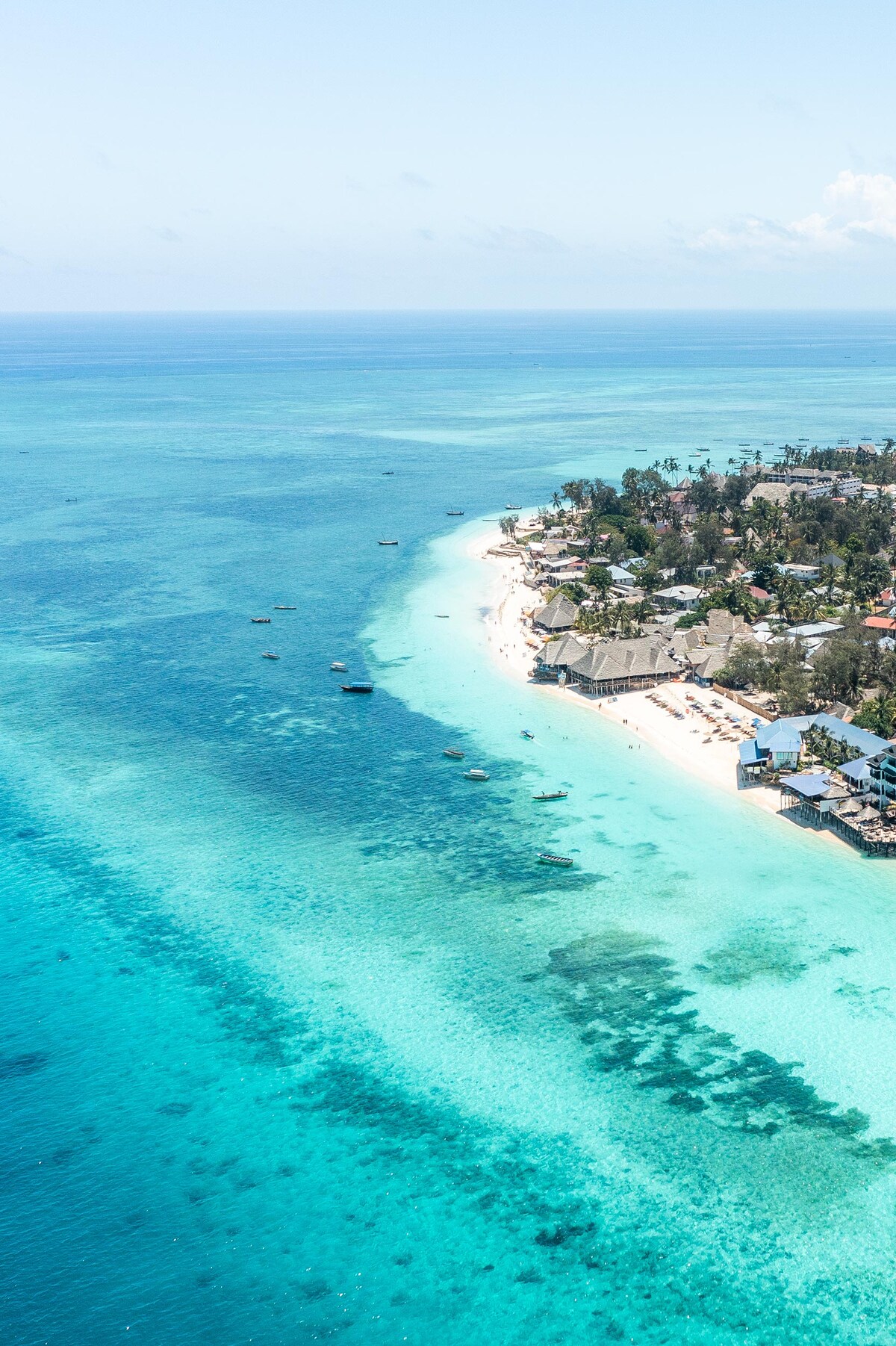 An aerial view showcases the stunning coastline of Nungwi beach, with clear turquoise waters gently lapping against the shore. The sandy beach is dotted with palm trees and beach umbrellas, while traditional boats can be seen in the distance on the vibrant sea.