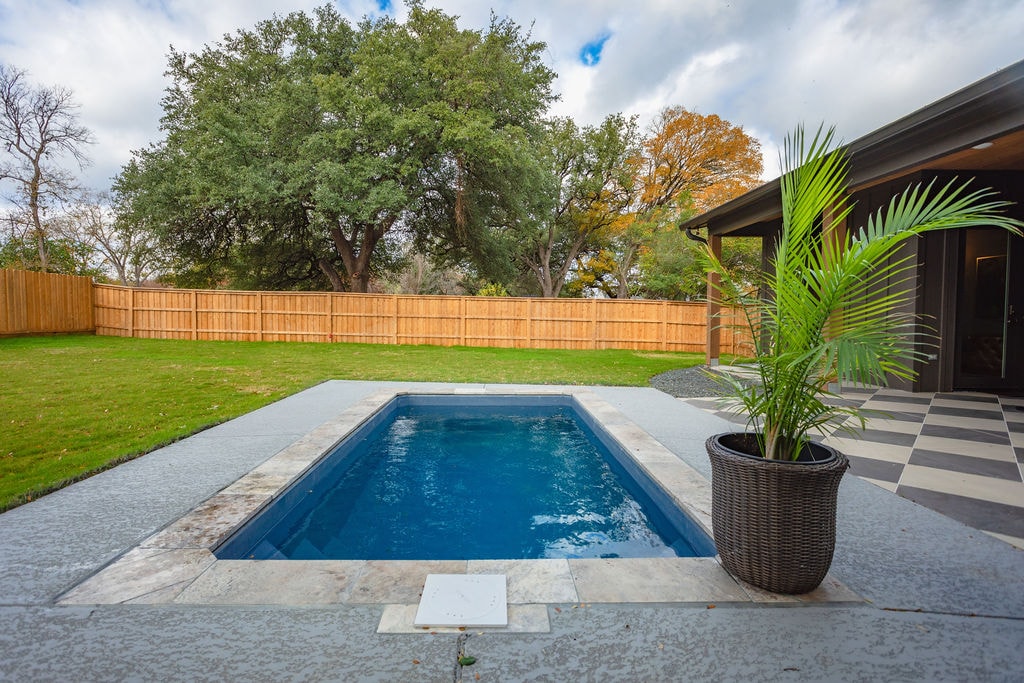 A small swimming pool is featured in the foreground, surrounded by a stone patio. Lush greenery is visible, including a large oak tree. A decorative potted plant adds a touch of vibrant green near the pool's edge, and a fenced backyard offers privacy.