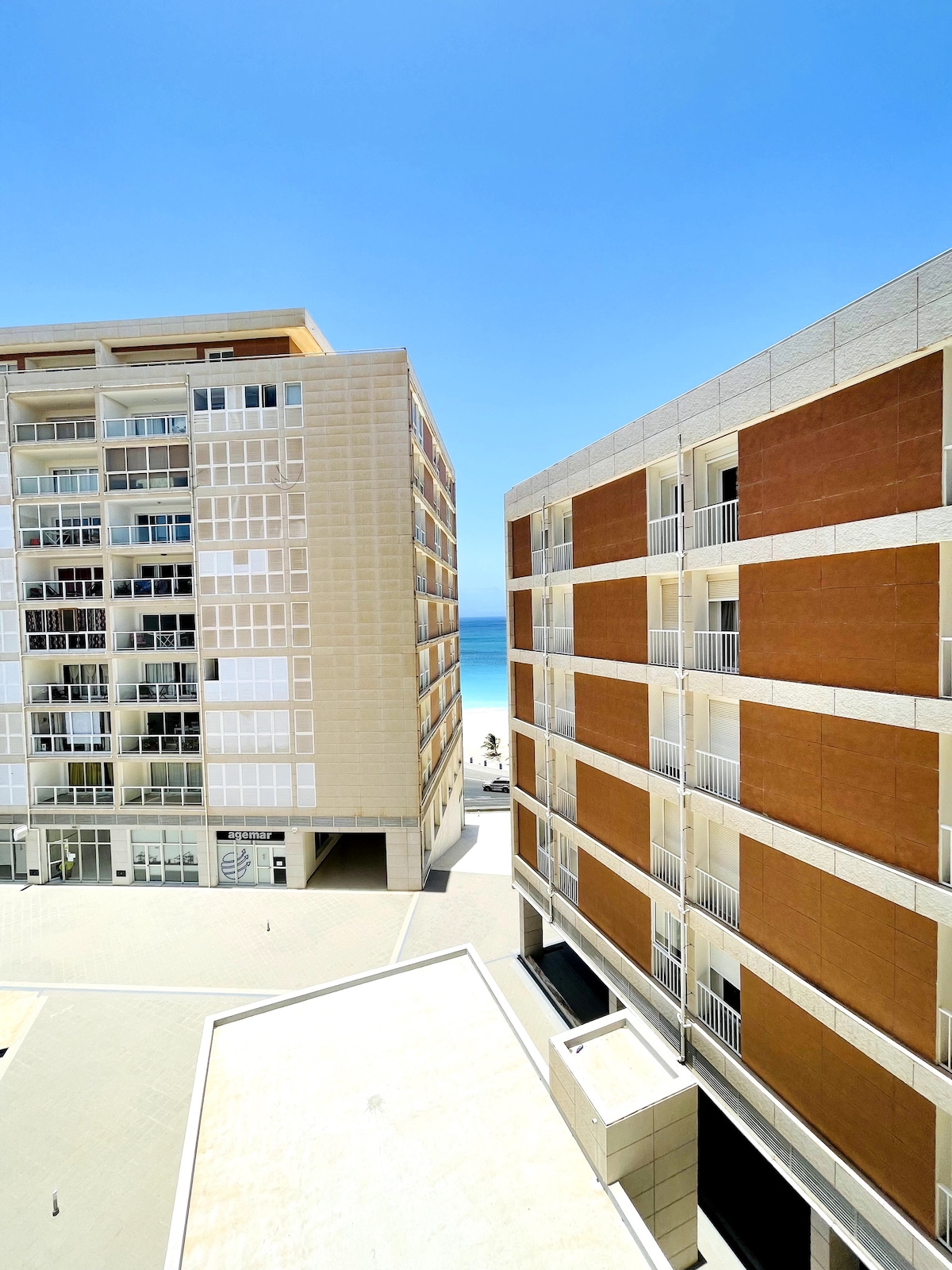 A view captures two modern apartment buildings with a combination of light-colored facades. The bright blue sky is visible above, and the ocean can be seen in the distance, suggesting proximity to the beach.