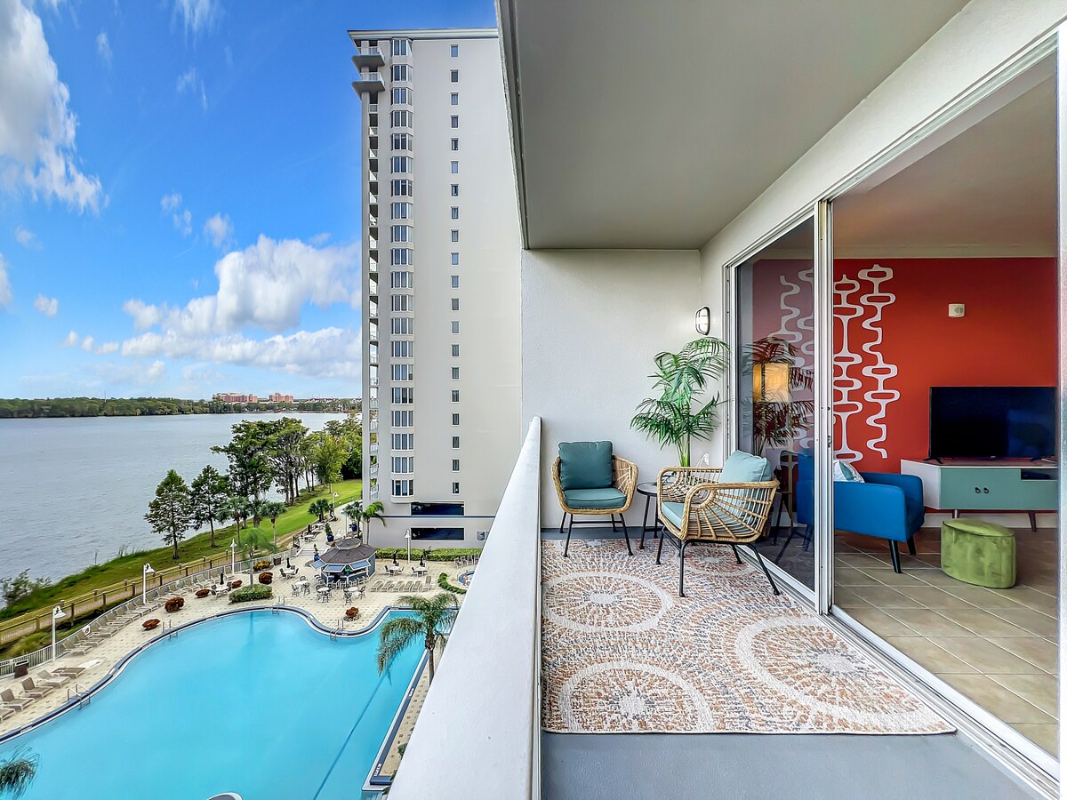 A private balcony overlooks a sparkling pool surrounded by lush greenery and a serene lake. Comfortable seating is arranged on the balcony, inviting relaxation while enjoying the scenic view. A colorful interior wall and large glass doors create a seamless connection to the living space.