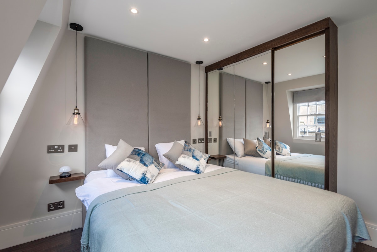 A cozy bedroom features a spacious bed with soft bedding and decorative pillows. Ambient lighting is provided by hanging fixtures on either side. A mirrored wardrobe reflects the room's layout, while natural light enters through a window with elegant window treatments.