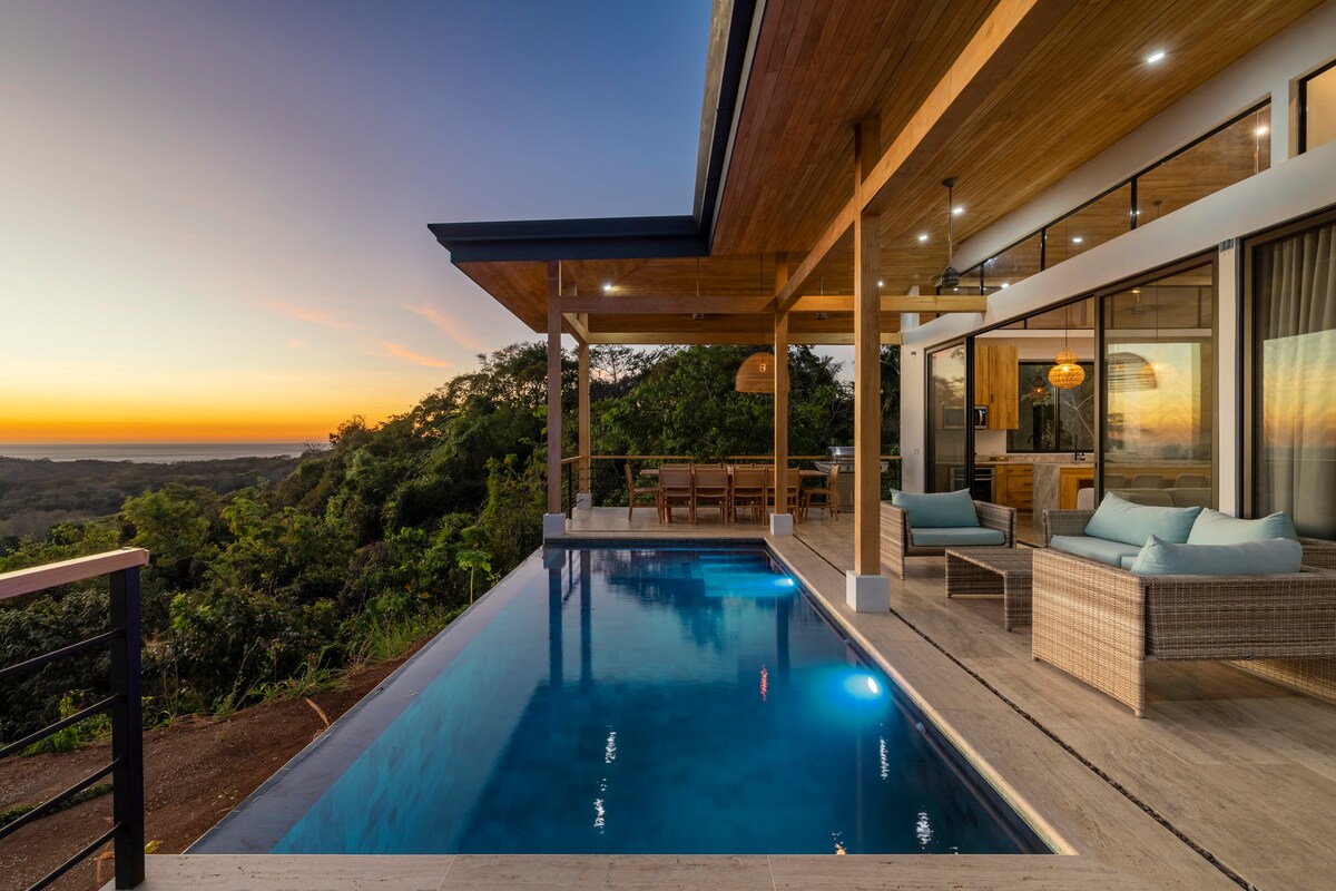 An expansive patio features a tranquil infinity saltwater pool, with wooden lounge furniture arranged nearby. The setting showcases lush greenery and ocean views in the background, illuminated by the soft glow of sunset. Natural materials and large windows connect the indoor and outdoor spaces.