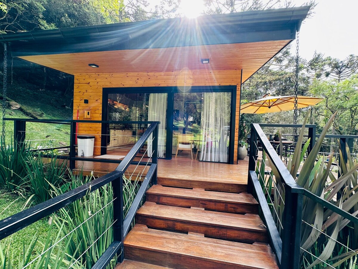 A welcoming entrance features a wooden deck leading to a bright, modern house surrounded by greenery. Large glass doors allow natural light to fill the interior, with a patio umbrella visible on the side, contributing to a serene outdoor setting.