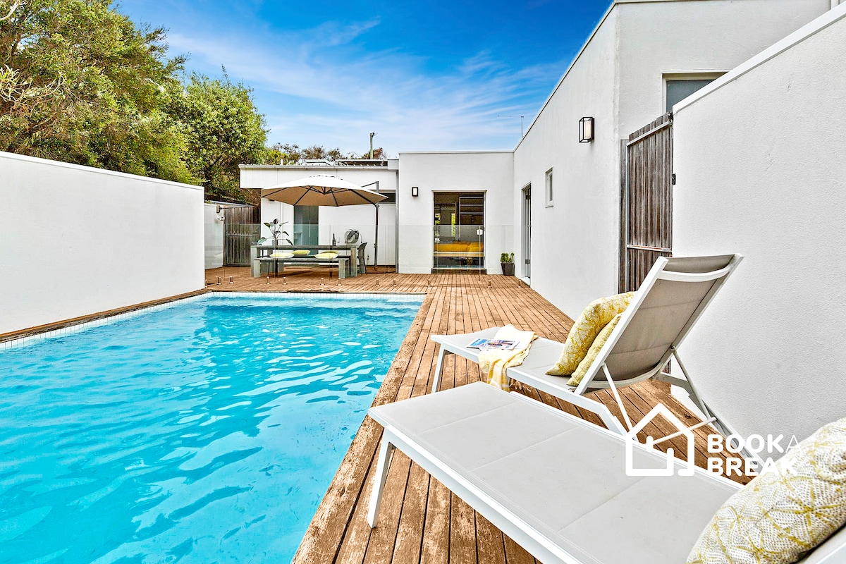A solar-heated pool is surrounded by a wooden deck, complemented by a seating area with an umbrella. Two lounge chairs are positioned poolside, inviting relaxation. The white-painted walls contribute to a bright atmosphere, with greenery visible in the background.