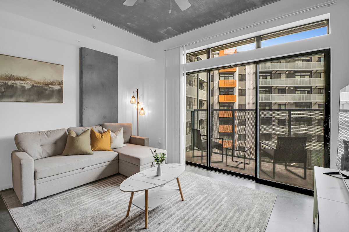A modern living room is presented with a comfortable sofa adorned with decorative pillows. A coffee table sits on a textured rug, and large sliding glass doors open to a balcony overlooking a city view. Soft lighting and minimal decor create an inviting ambiance.