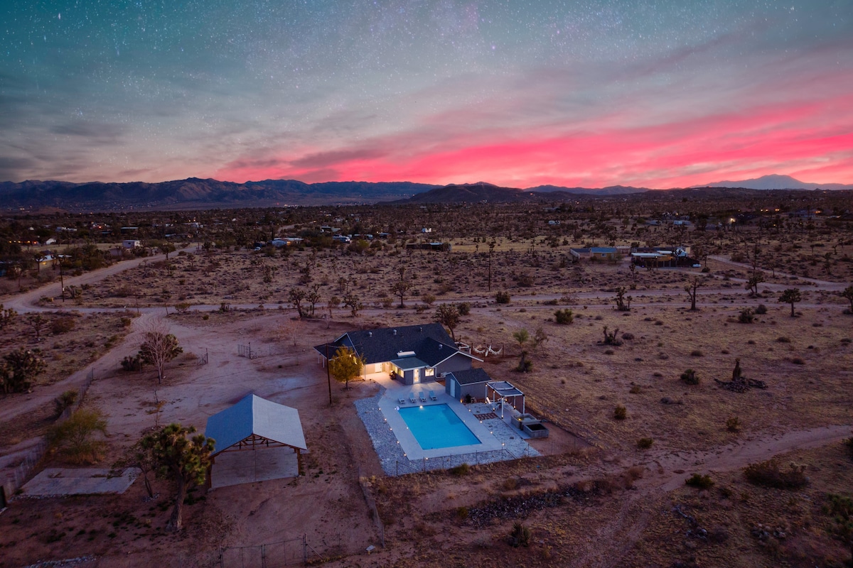 A spacious property set against a vibrant sunset sky, showcasing a private Olympic-sized swimming pool surrounded by a contrasting sandy landscape. The home is illuminated warmly, with desert vegetation peppering the area, extending towards distant mountain outlines.