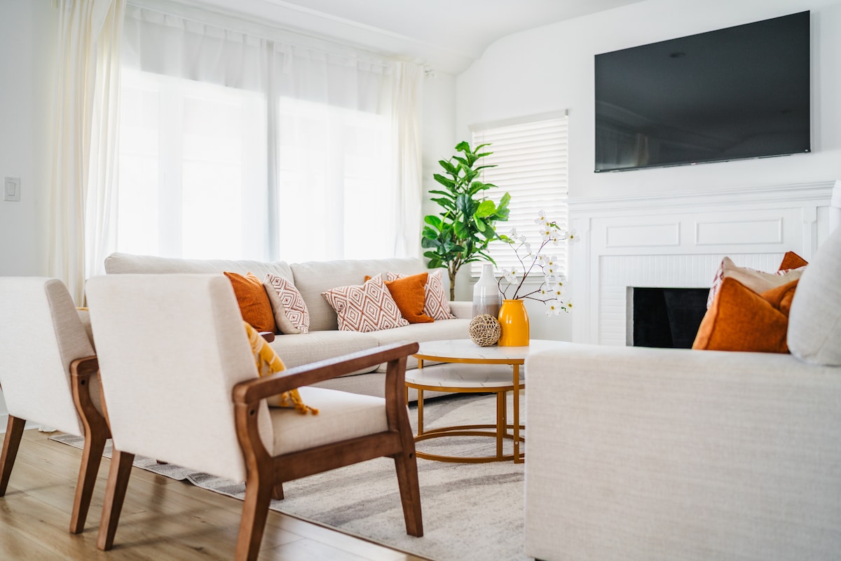 The living area features a spacious arrangement of light-colored sofas and armchairs with accent pillows. A stylish coffee table sits in the center, complemented by a decorative vase. Natural light filters through sheer curtains, enhancing the relaxed atmosphere.