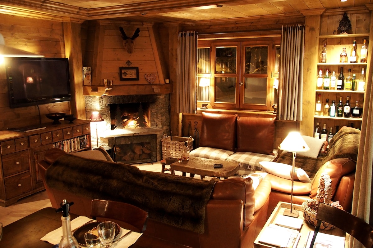 A warm living area is depicted, featuring a large leather sectional sofa arranged around a stone fireplace. A wooden entertainment unit holds a flat-screen television, and shelves display an assortment of bottles. Soft lighting enhances the cozy atmosphere.