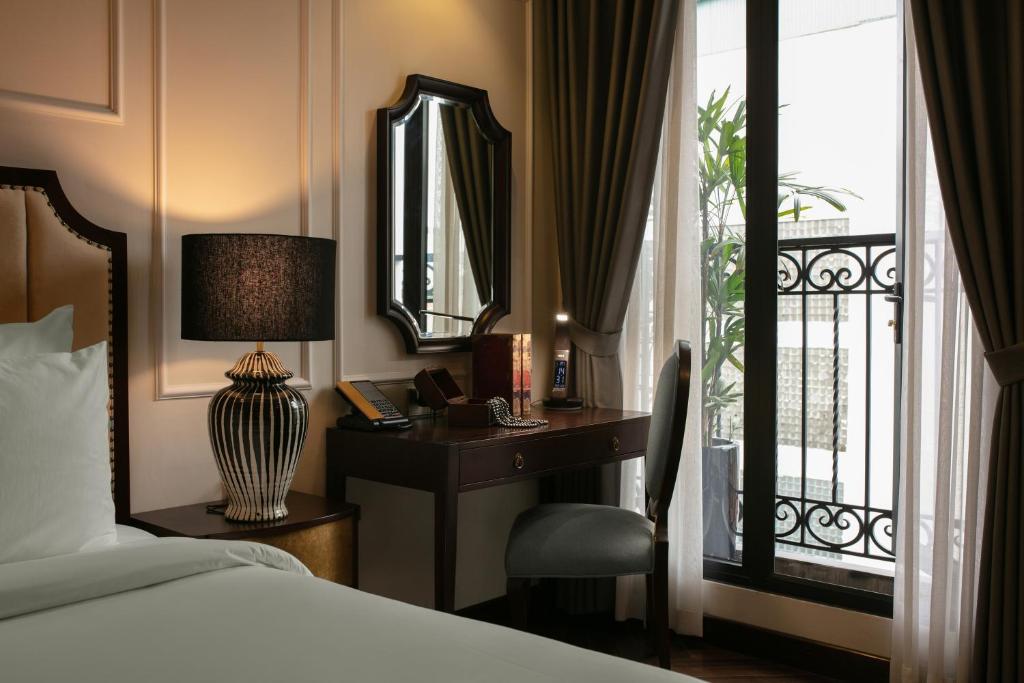 A cozy room features a spacious bed with crisp white linens, complemented by a decorative lamp on a stylish bedside table. A writing desk with a mirror is positioned near a window that offers views of an outdoor plant, framed by elegant curtains.