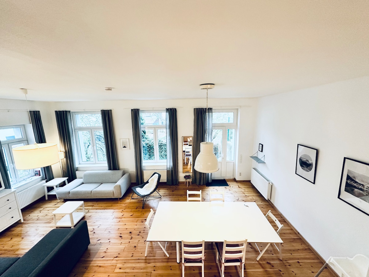 A spacious living area captures natural light through large windows, complemented by tall gray curtains. A light wooden dining table is surrounded by white chairs, while a comfortable seating arrangement features a gray sofa and a lounge chair in the corner. A minimalist pendant light hangs above the table.