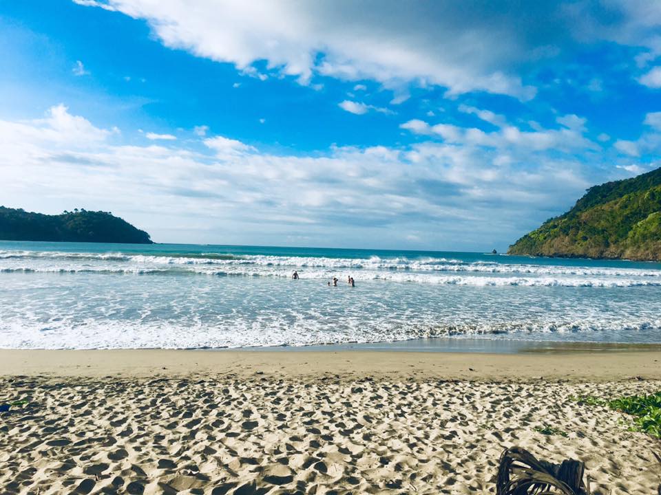 A sandy beach stretches into the distance, bordered by gentle waves lapping at the shore. In the background, lush green hills rise against a bright blue sky dotted with clouds. The scene captures people enjoying the water, enhancing the tranquil beach atmosphere.