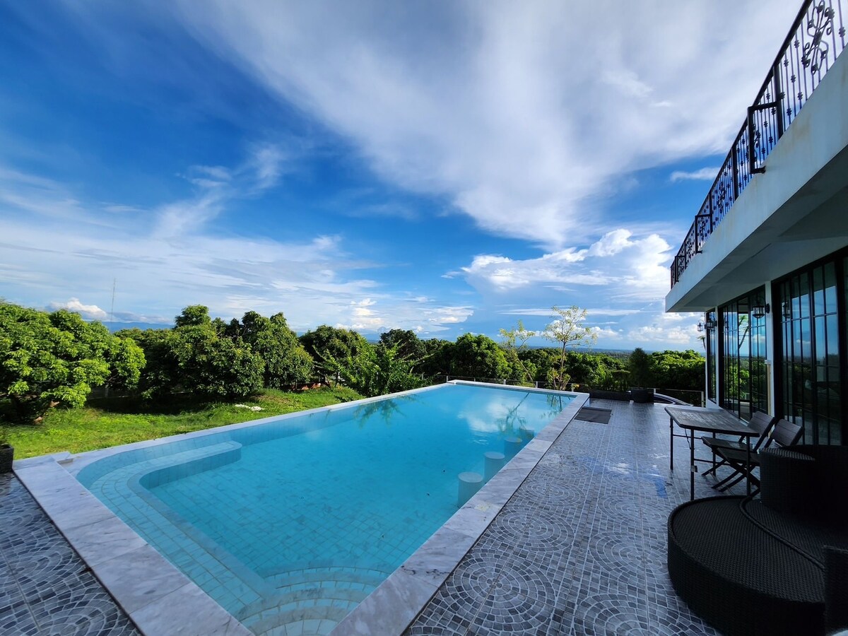 A spacious private pool is seen set against a backdrop of lush greenery and open skies. The pool area is bordered by elegant stonework, providing a serene environment for relaxation. A dining set can be spotted on the deck, with large glass doors leading to the villa.