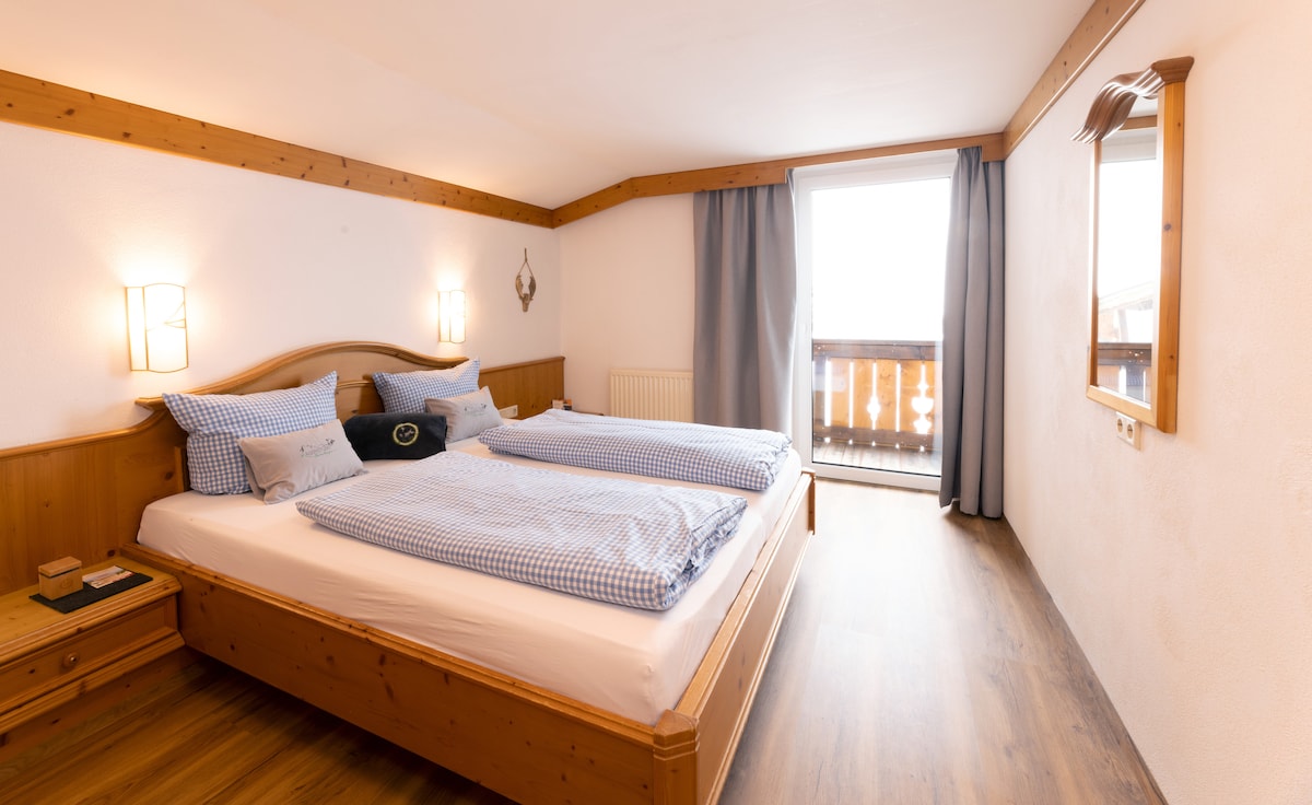 A well-appointed bedroom features two spacious beds dressed in blue checkered linens. Natural light enters through the balcony door, which leads to a small outdoor space. Wooden accents and soft lighting create a calming ambiance.