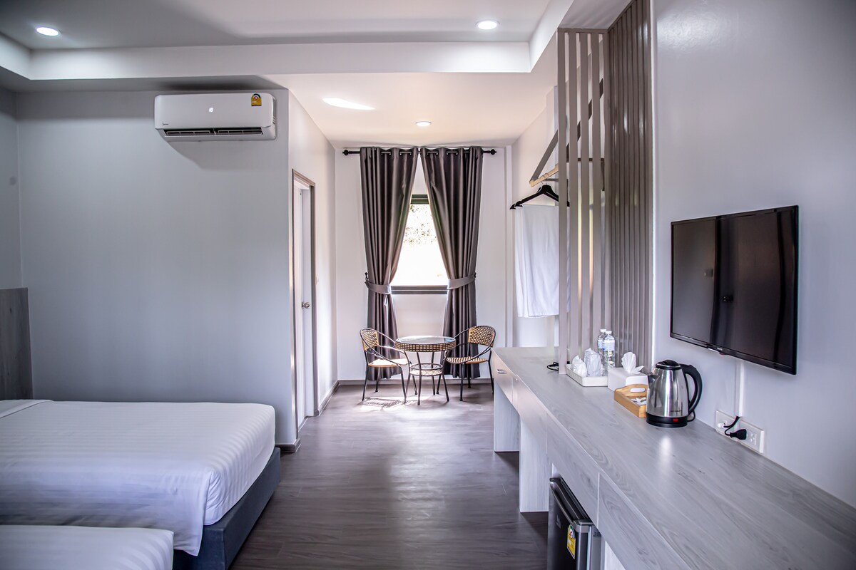 A spacious room features two single beds dressed in crisp white linens. Natural light fills the area through large windows, complemented by gray curtains. A small table with two chairs is positioned near the window, while a flat-screen TV and kettle are neatly arranged on a console.