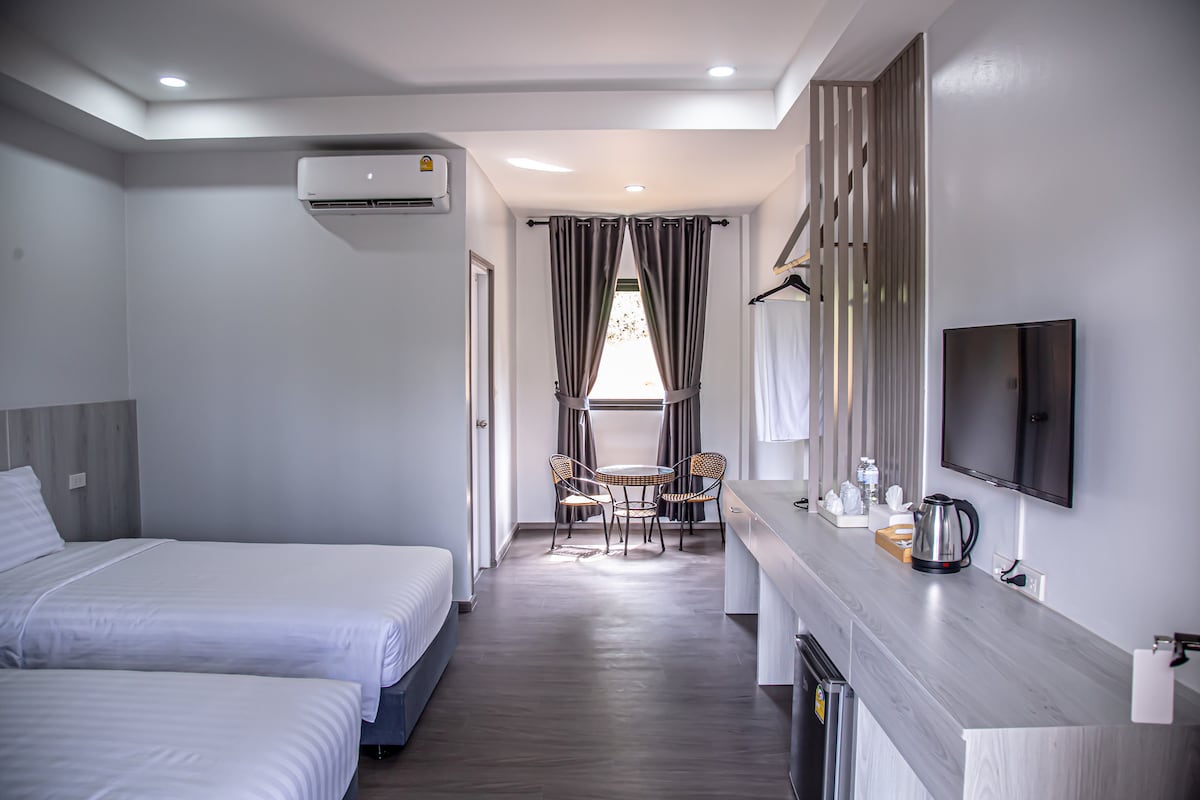 A modern room is shown with two single beds dressed in white linens. A small table with two chairs is positioned near a window dressed with dark curtains. An entertainment unit with a television, kettle, and complimentary items is centered against a light grey wall.