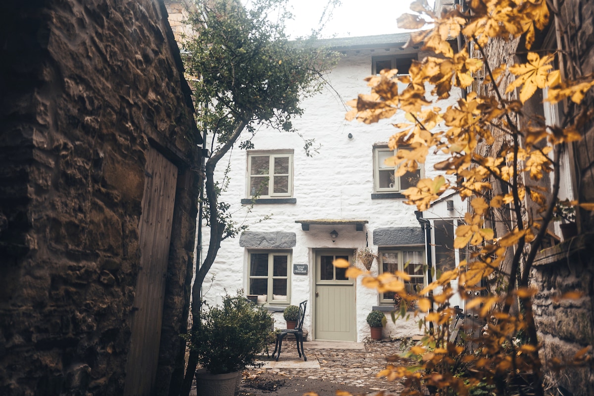 A charming cottage exterior is framed by lush foliage and rustic stone walls. A small seating area is visible beside the entrance, inviting relaxation. The soft light highlights the textured white facade, complemented by surrounding autumn leaves, showcasing the cottage's character and historical essence.