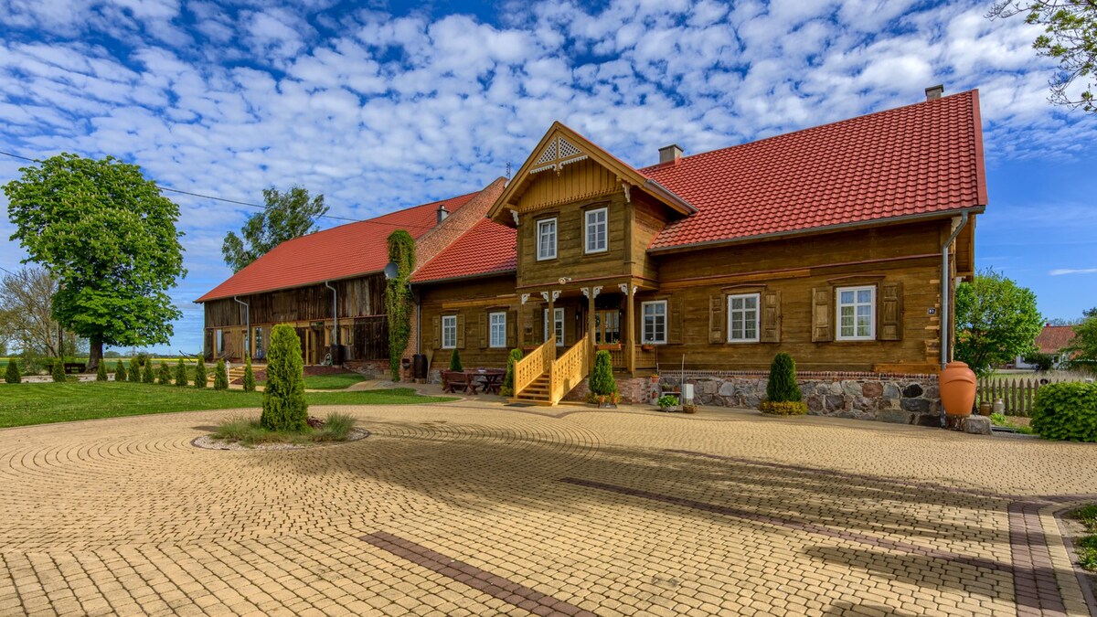 A spacious wooden house is set against a backdrop of blue skies, with a distinctive red roof and neatly arranged stone pathways. Lush greenery surrounds the property, enhancing the cozy, rural setting. A welcoming entrance is highlighted by steps leading to a wooden porch.