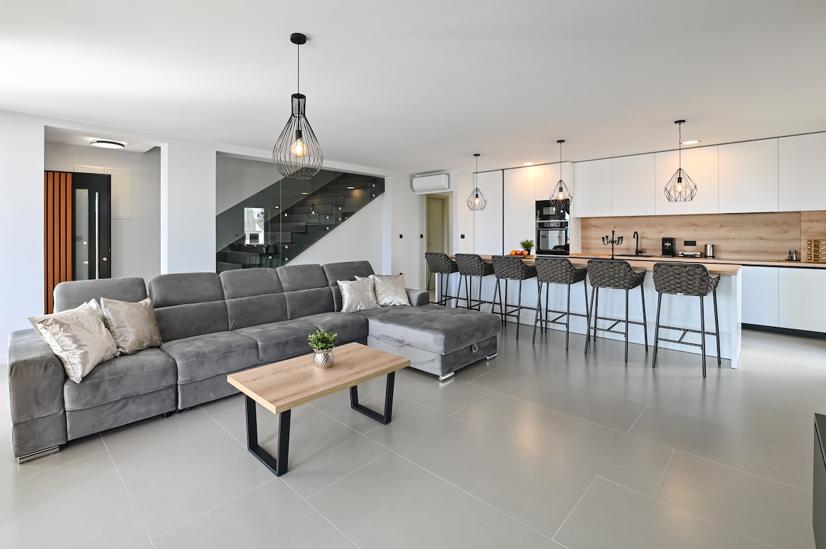 A spacious living area features a grey sectional sofa complemented by a wooden coffee table. A modern kitchen with bar seating is visible in the background, illuminated by pendant lighting. Light-colored walls and tiled flooring enhance the open and contemporary design.
