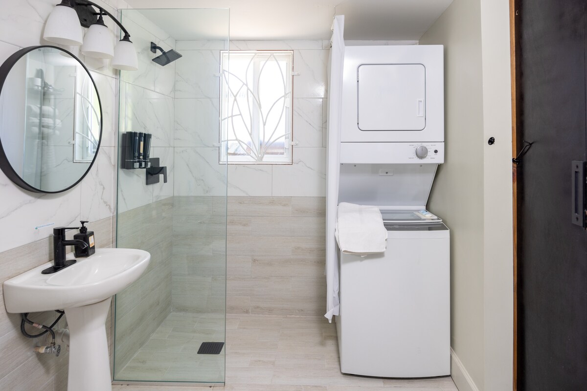 A modern bathroom is featured, showcasing a glass-enclosed shower with a rainfall showerhead and elegant tiled walls. A sleek circular mirror complements a compact sink. A washer and dryer unit is positioned in the corner, providing convenient laundry access.