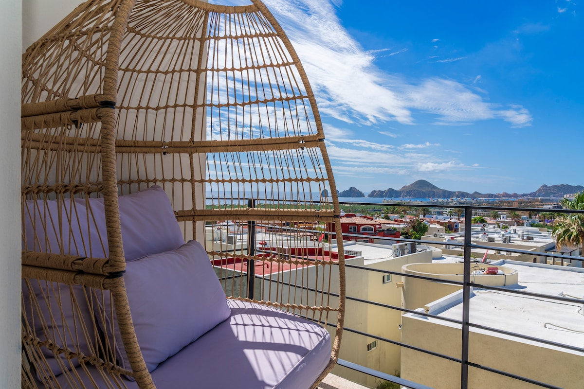 A rattan hanging chair offers a comfortable seating area on the private terrace. The stunning ocean view displays the Sea of Cortez, with rocky formations in the distance and a clear blue sky above. Soft cushions enhance the space for relaxation.