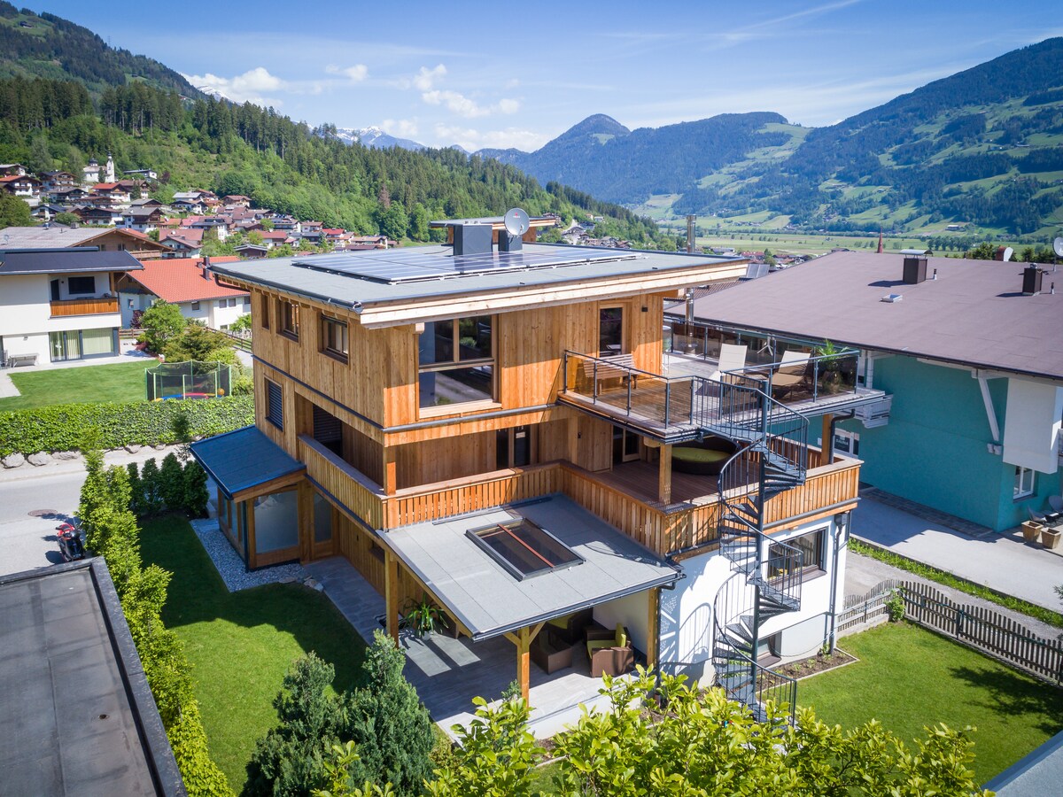 The exterior of the Spieljochlodge Penthouse presents a modern wooden structure set against a backdrop of the Tiroler Alps. Multiple balconies and a spiral staircase are visible, offering access to the upper levels. Lush greenery surrounds the property, enhancing the inviting atmosphere.