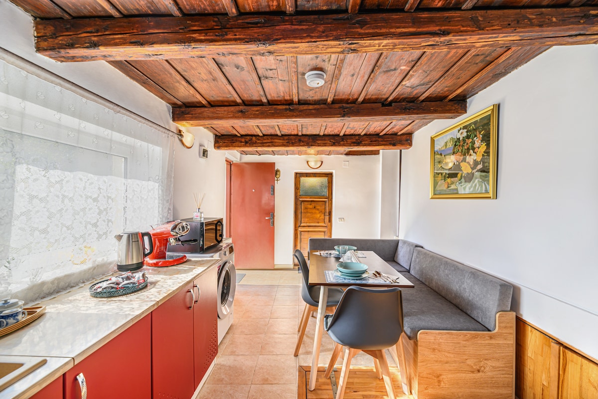 A well-appointed kitchen area features exposed wooden beams and a spacious countertop. Modern appliances, including a kettle and microwave, are visible. A dining table is accompanied by comfortable seating, while natural light filters through a lace curtain, creating an inviting space.