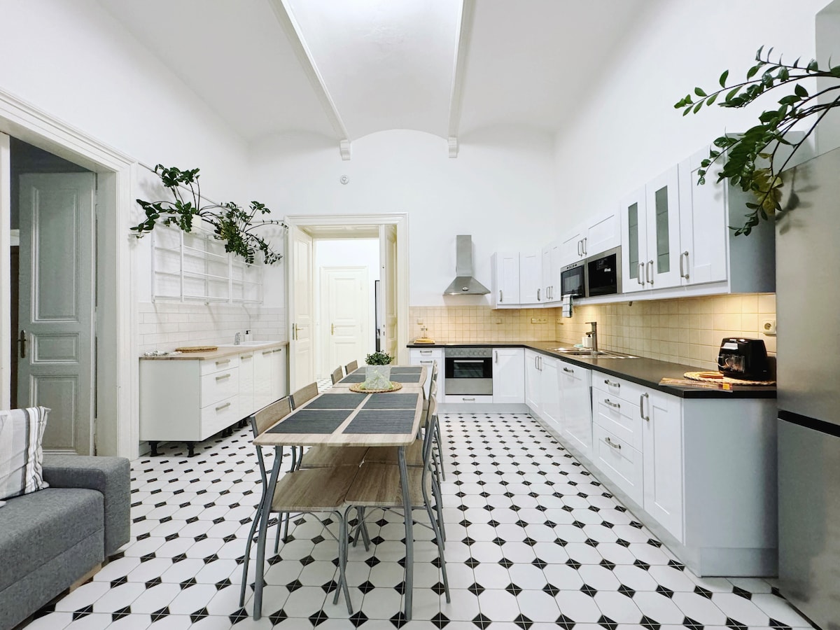 A spacious kitchen features sleek white cabinetry and modern appliances, illuminated by natural light from the skylight. A long wooden dining table is surrounded by metal chairs, and greenery decorates the walls, enhancing the inviting atmosphere.