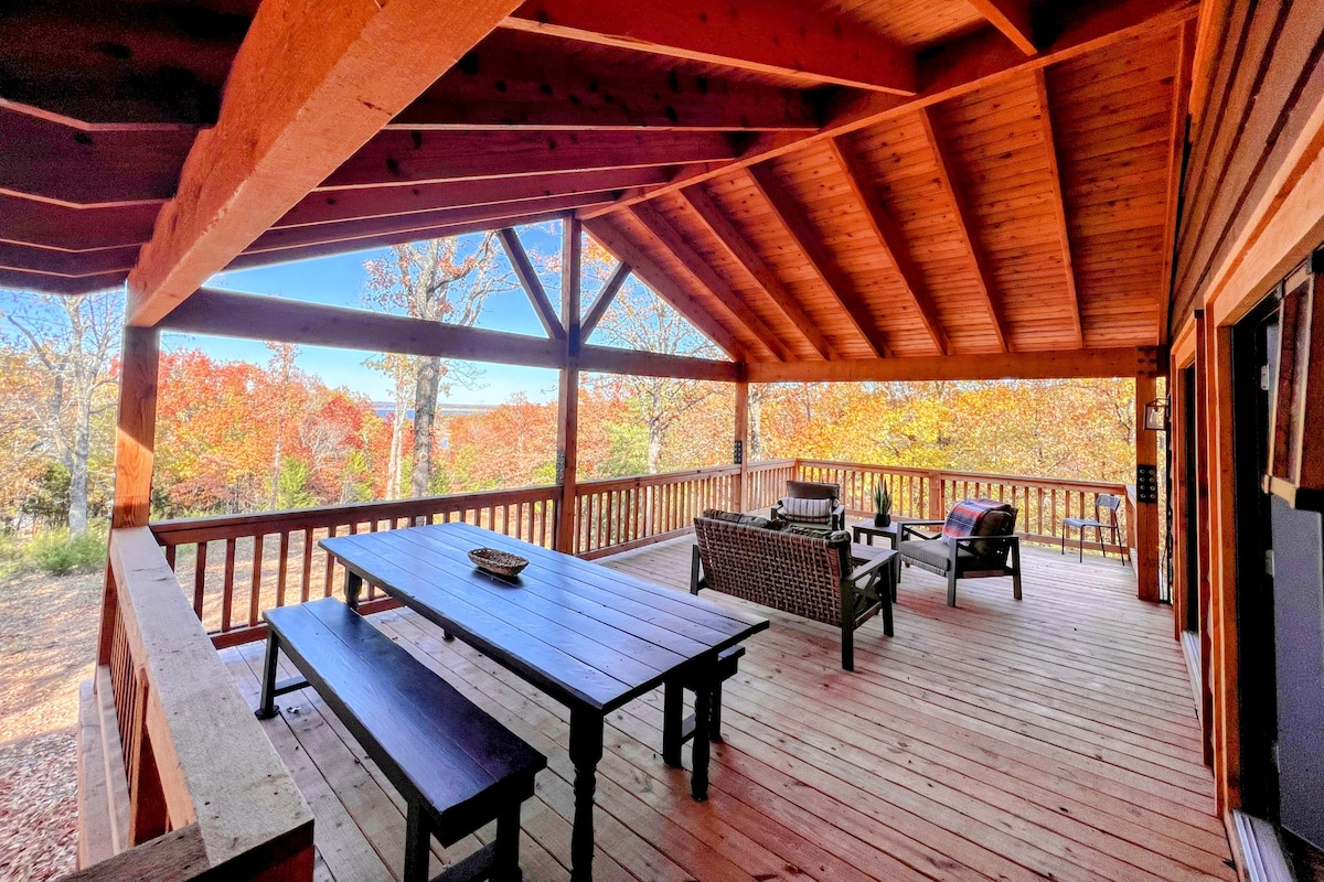 A covered cedar deck provides ample seating and dining space, with a long wooden table and comfortable chairs. The surroundings are filled with vibrant fall foliage, enhancing the sense of nature. Wooden beams support the high ceiling, while large openings invite fresh air and views of the outdoors.