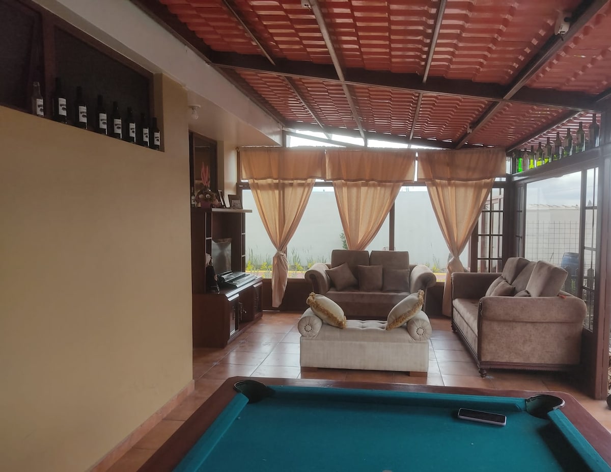 A spacious game room is highlighted by a billiards table centered in the area. Comfortable seating is arranged with a beige sofa and an armchair adorned with cushions. Large windows draped with light curtains allow natural light, providing an airy feel to the space.