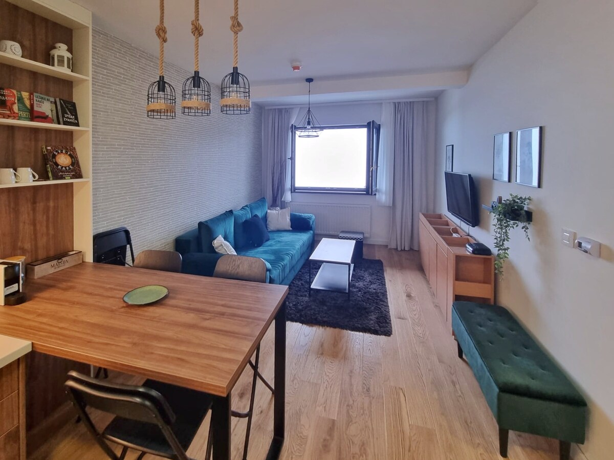 A modern living area is furnished with a plush teal sofa, a coffee table, and a wooden dining table. A flat-screen TV is mounted on the light-colored wall. Natural light enters through a large window draped with soft curtains, providing a warm atmosphere.