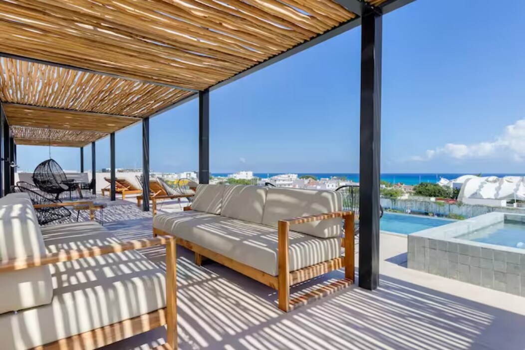 An outdoor seating area is showcased under a wooden pergola, offering a view of the ocean. Comfortable sofas and lounge chairs are arranged on a spacious deck, with sunlight creating a pattern across the surface. A swimming pool is visible in the background.