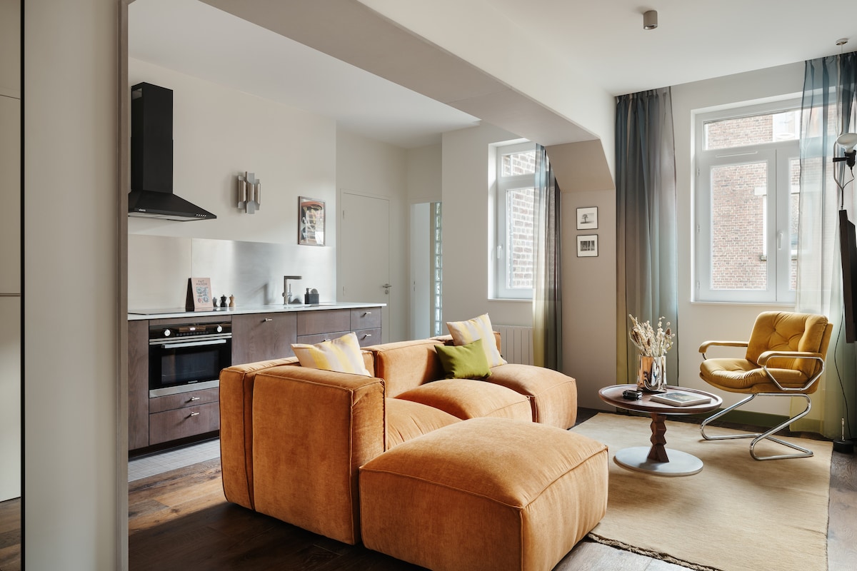 An inviting living area features a modern, curved orange sofa with accent pillows, paired with a round coffee table. Light streams in through large windows adorned with sheer curtains, illuminating the space. The kitchen area is visible in the background, equipped with contemporary appliances.