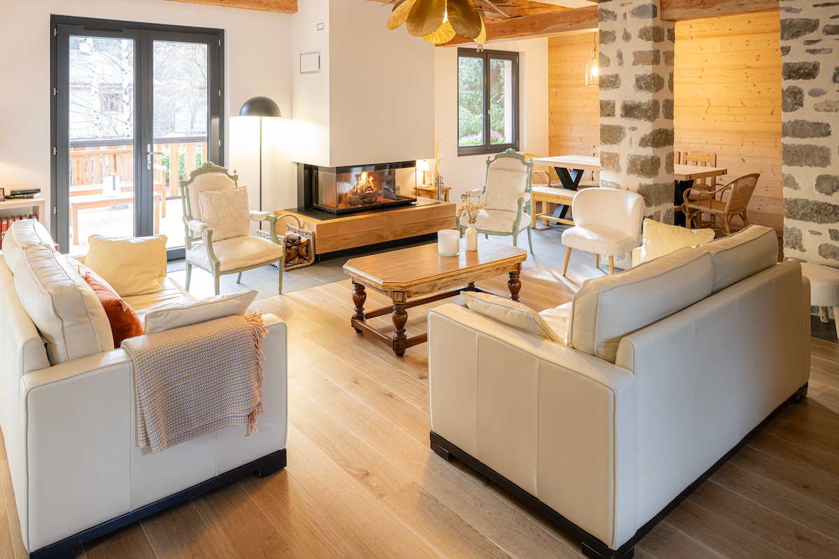A spacious living area features two light-toned leather sofas facing a wooden coffee table. Natural light fills the room through large sliding glass doors that lead to a terrace. A cozy fireplace offers warmth, and wooden beams enhance the rustic charm of the space.