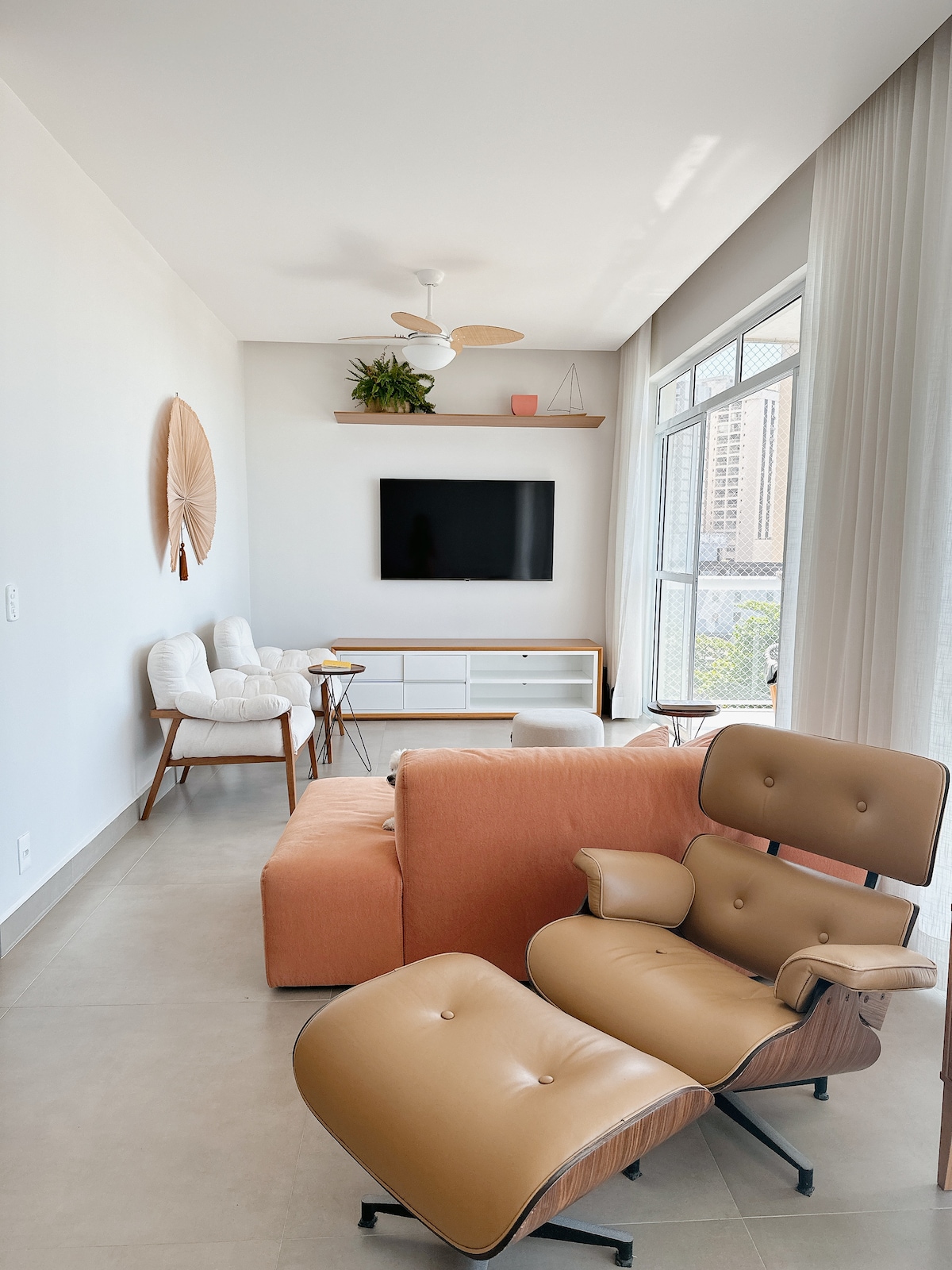 A bright living area is presented, featuring a vibrant orange sofa and a contemporary beige chair. Large windows allow natural light to fill the space. A mounted television and light wooden shelving are visible, with greenery and decorative items adding a touch of warmth.