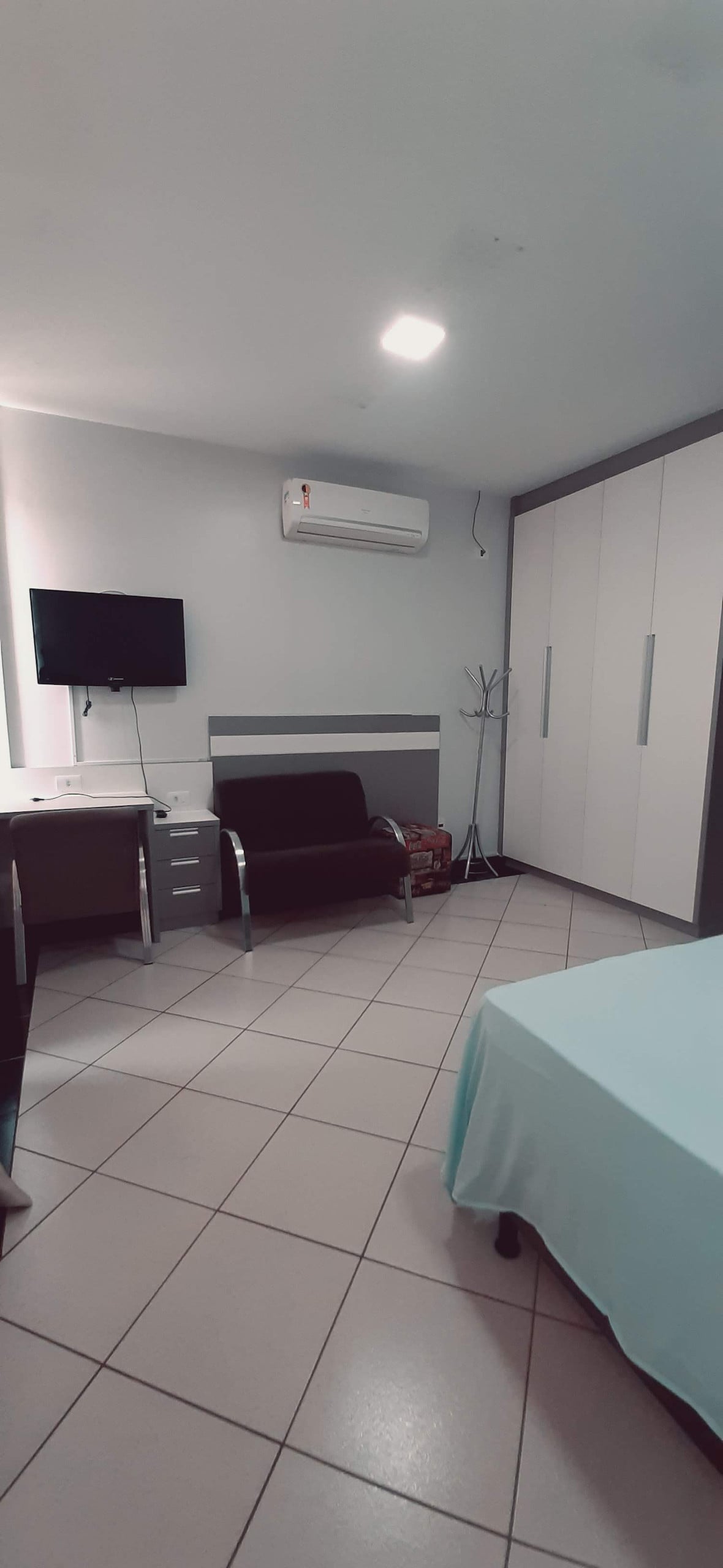A spacious bedroom is equipped with a large wardrobe, desk with a chair, and a black seating area. A television is mounted on a wall. Soft lighting enhances the clean lines of the furnishings, while a cool color palette contributes to the room's airy feel.