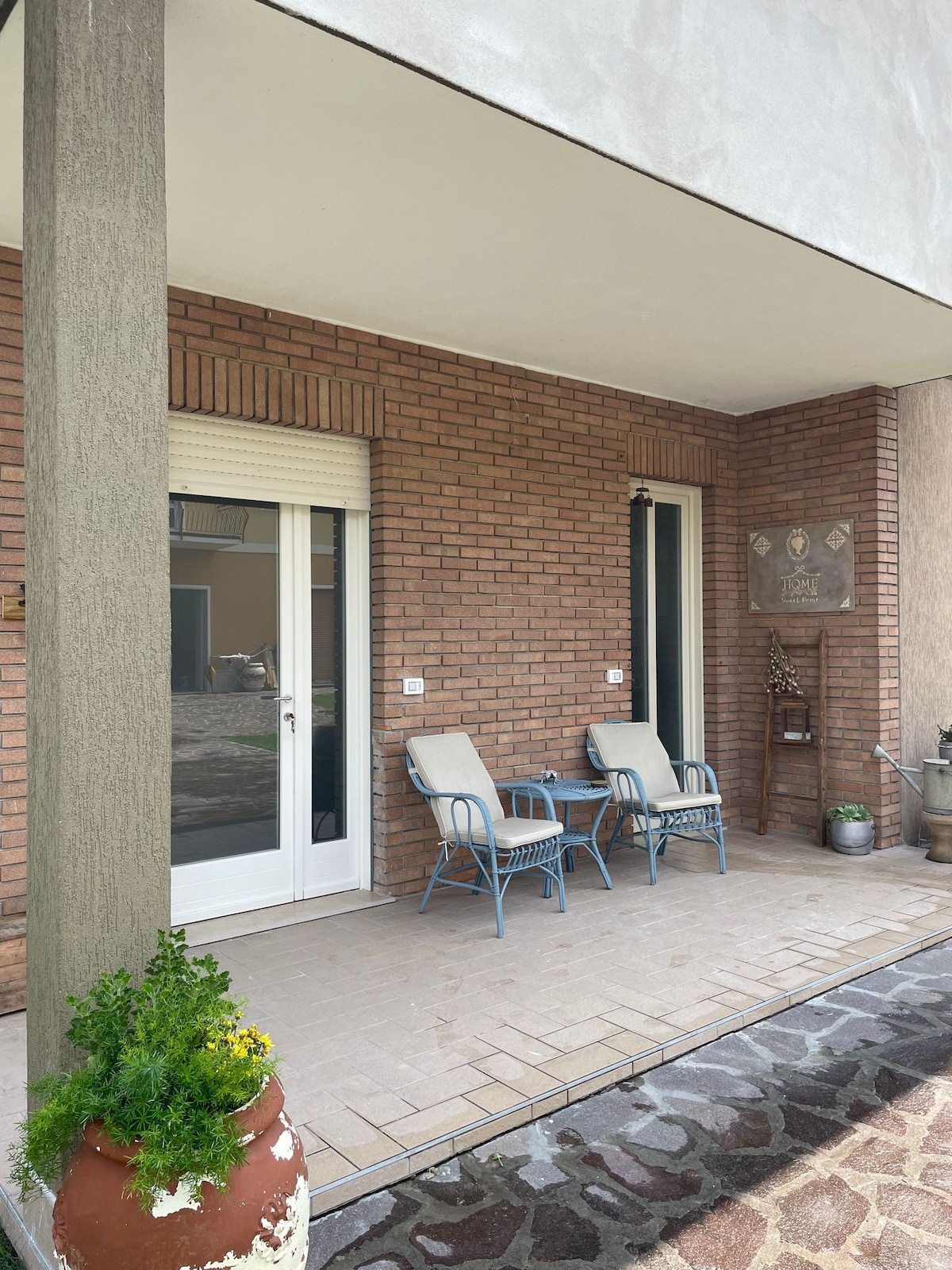 A small porch area features two blue chairs positioned on a tiled floor, adjacent to a light-colored door. Decorative elements, including a plant, add character to the space, which is framed by a stylish brick wall.
