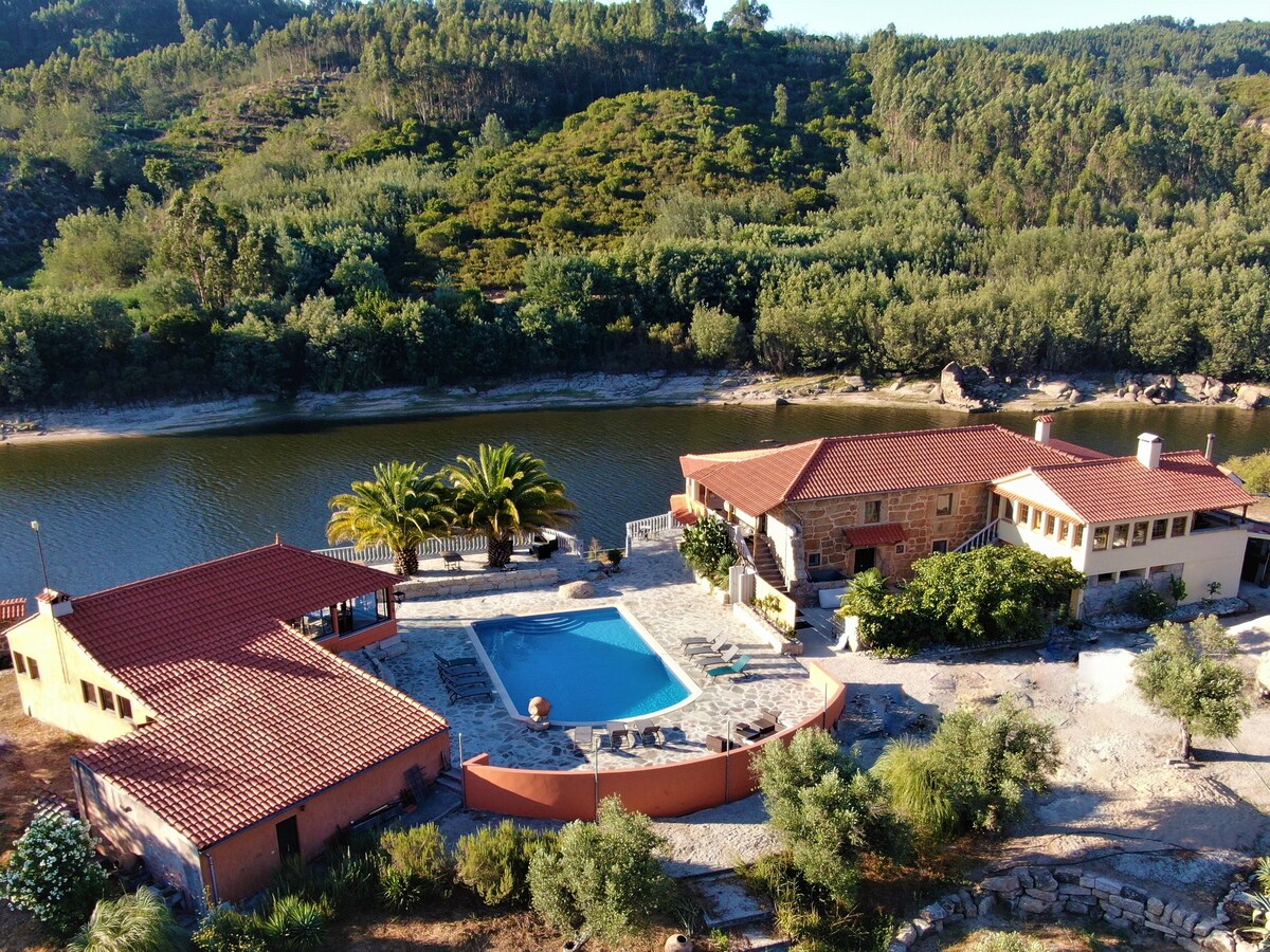 An aerial view captures a tranquil property along the Rio Mondego. A clear blue pool is surrounded by a sun terrace with loungers, while the adjacent stone building features red tiled roofing. Lush greenery and palm trees frame the landscape, complementing the serene waterway.