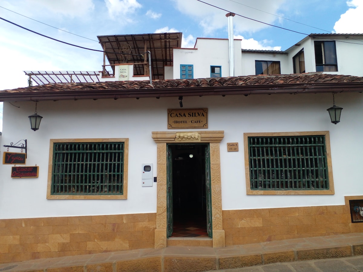 The exterior of a charming hotel is presented, featuring white walls and a tiled roof. Large windows with green metal bars provide character. A decorative sign indicates 'Casa Silva Hotel-Café,' while a light-colored stone foundation adds a sturdy element. The entrance is centrally located.