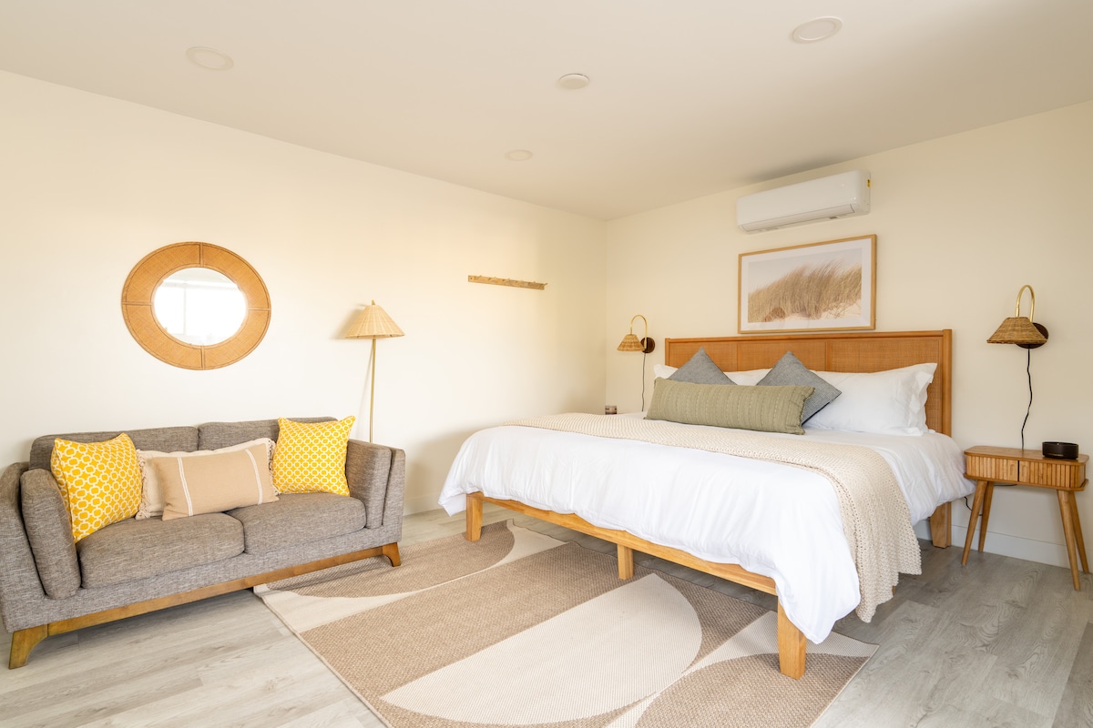 A bright and airy studio features a spacious king-size bed adorned with layered pillows and a textured throw. A cozy sofa with yellow accent cushions complements the neutral decor. Natural light bathes the room, highlighting the round mirror and stylish accents throughout.