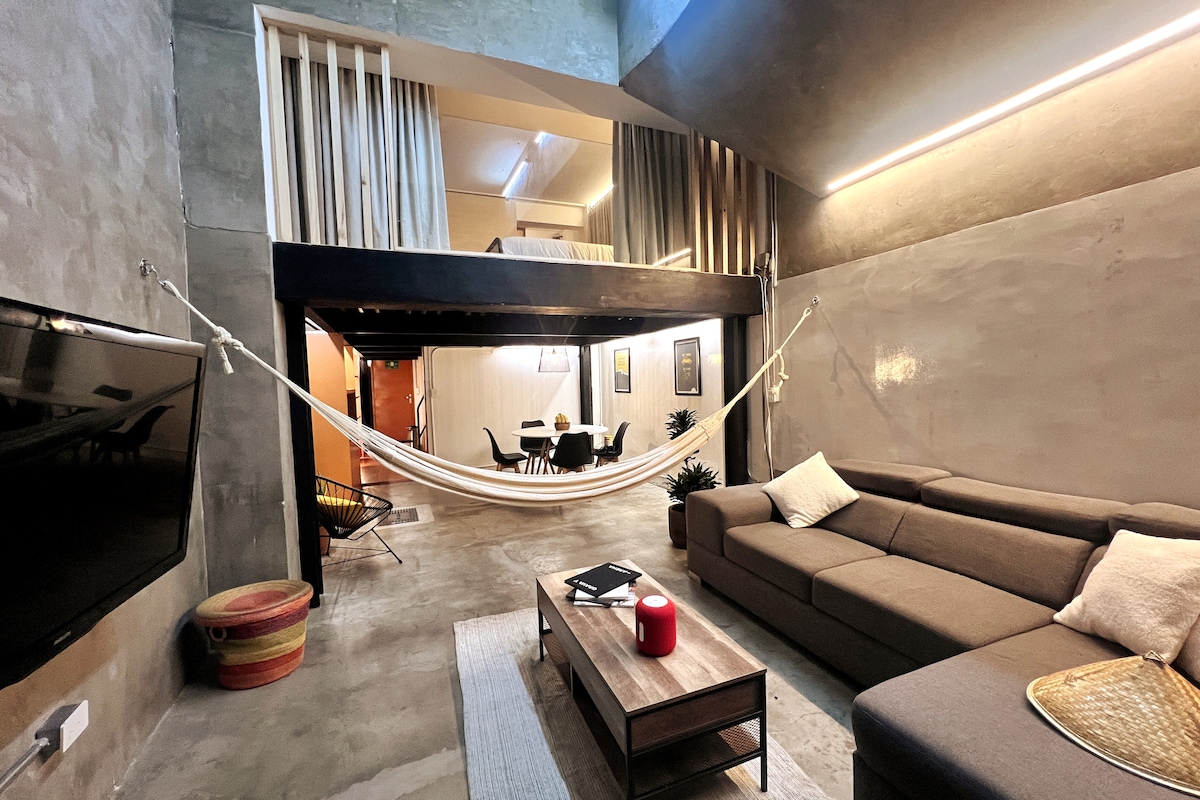 An open living space features a large, gray sectional sofa positioned near a low coffee table. A hammock is gently suspended above, creating a relaxed atmosphere. A dining area with a round table and chairs can be seen in the mezzanine. The industrial-style walls are finished in polished concrete.