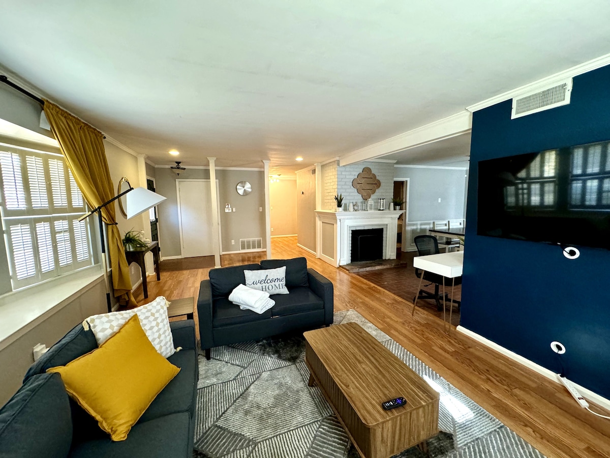 A spacious living area is featured, showcasing a comfortable gray sofa accented with yellow and patterned pillows. A large coffee table sits on a patterned rug, and a flat-screen TV is mounted on a vibrant blue wall. Natural light enters through the window with stylish shutters.