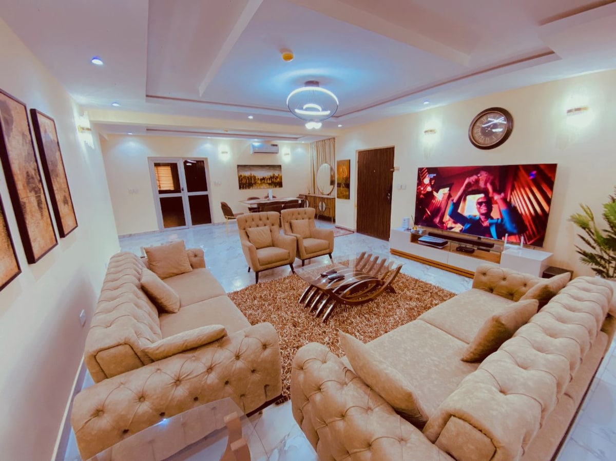 A spacious living area is furnished with a comfortable arrangement of plush sofas and armchairs, creating a welcoming ambiance. A large television is mounted on the wall, displaying entertainment, while a coffee table sits at the center on a textured rug. Soft lighting enhances the room's inviting feel.
