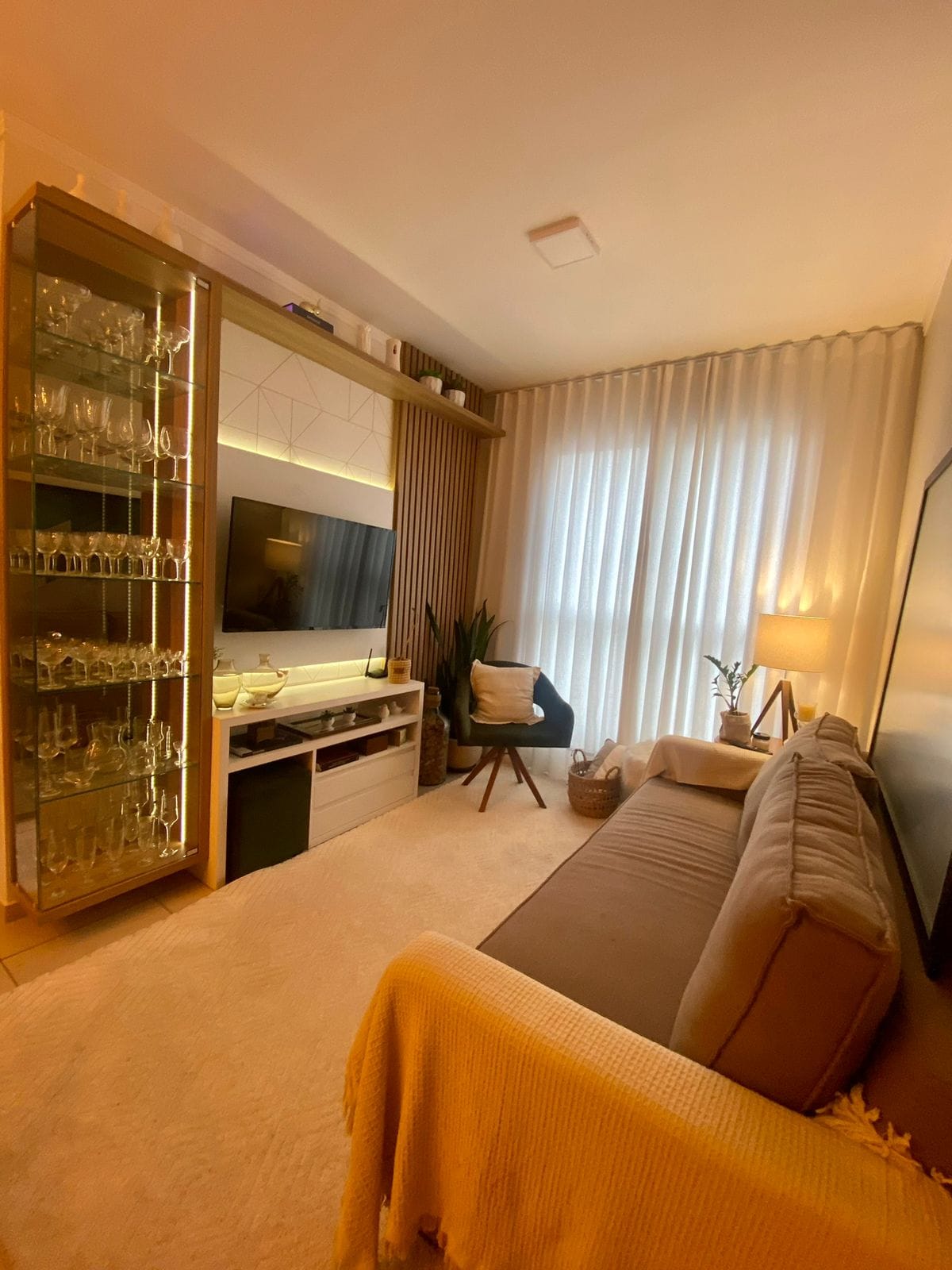 A cozy living area is featured, showcasing a comfortable sofa and a large wall-mounted television. Glass shelves display various decorative items. Natural light enters through sheer curtains, and a floor lamp adds a soft glow, enhancing the warm ambiance of the space.