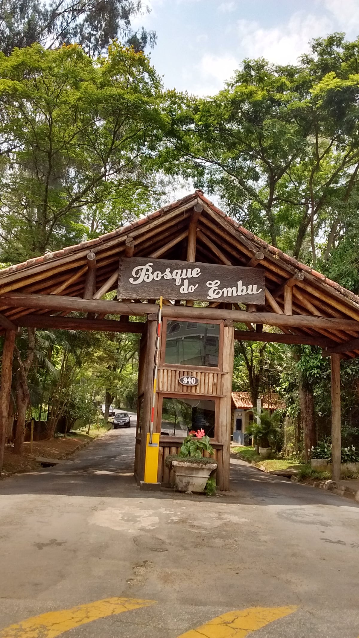 The entrance to Bosque do Emhu features a rustic wooden structure with a sloped roof, displaying the name prominently. Lush greenery surrounds the entrance, contributing to the natural setting. A pavement leads into the property, with a small decorative plant placed beneath the sign.
