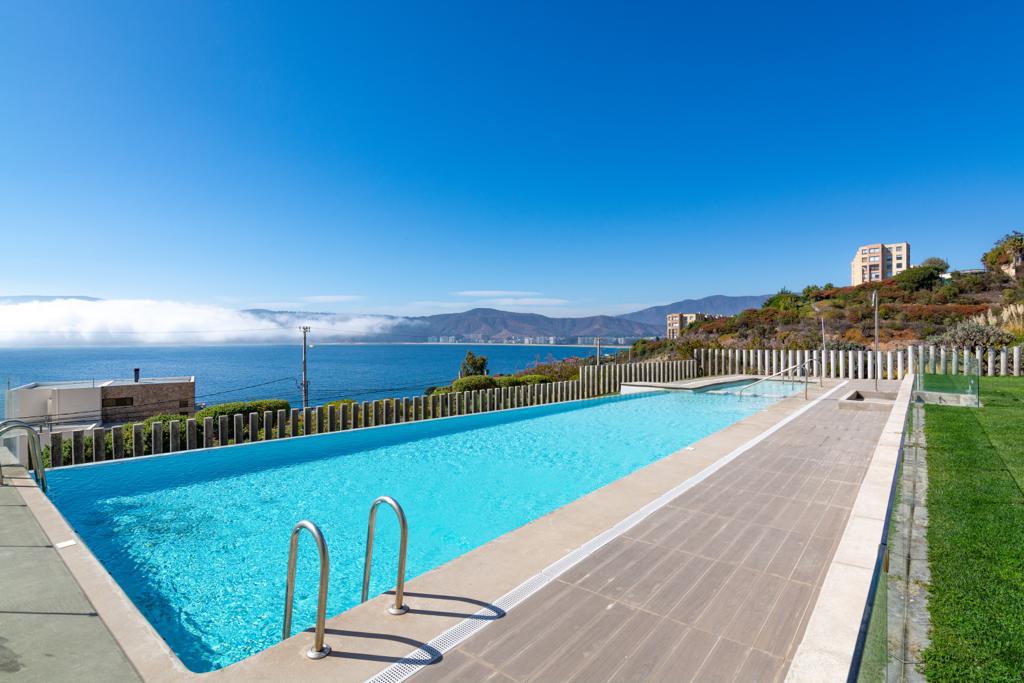 An outdoor swimming pool is visible, surrounded by a spacious deck and green grass. The clear blue water reflects the sky, while the tranquil sea and distant mountains create a serene backdrop. The area is well-maintained, with a railing along the pool's edge.